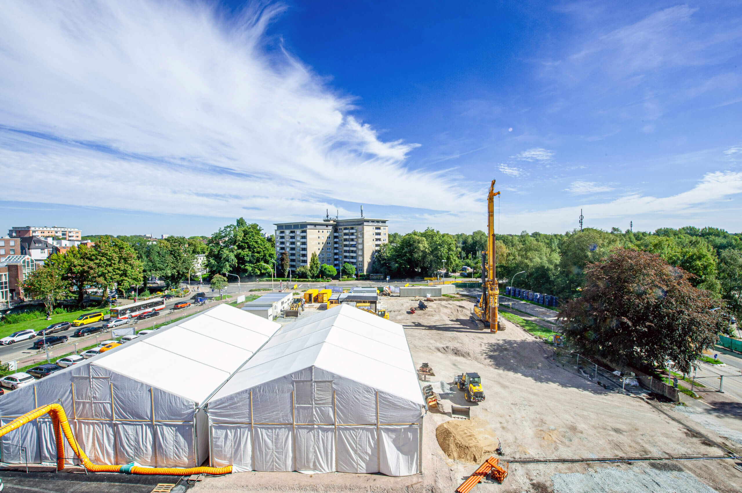 Baustelle eines innerstädtischen Entwicklungsprojekts mit großen weißen Schutzzelten, Baugeräten und Bohrgerät; im Hintergrund Wohngebäude, Straßenverkehr und Grünflächen unter blauem Himmel.
