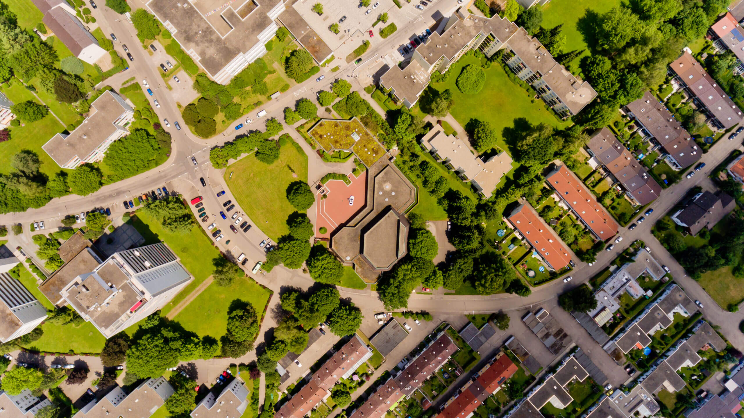 Luftaufnahme eines innerstädtischen Wohn- und Geschäftsquartiers mit Straßenkreuzung, begrünten Innenhöfen, Parkflächen und dicht angeordneten Gebäuden inmitten vieler Bäume.
