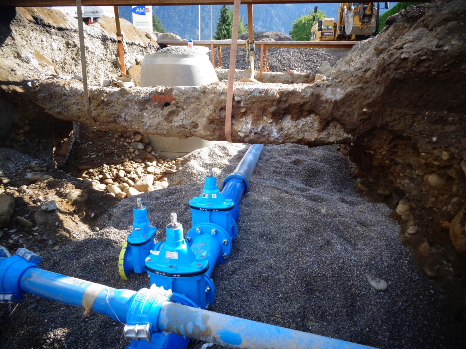 Neu verlegte blaue Wasserleitungen mit Absperrarmaturen in einer Baugrube; die Rohre liegen im Kiesbett, darüber eine freigelegte Erdschicht und Baumaschinen im Hintergrund.