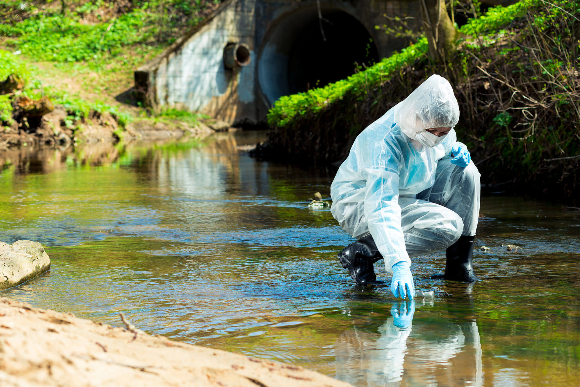 Fachkraft in Schutzanzug, Handschuhen und Atemschutz entnimmt eine Wasserprobe aus einem Bach; im Hintergrund ein Durchlassrohr, Sinnbild für Umweltuntersuchung und Gewässermonitoring.